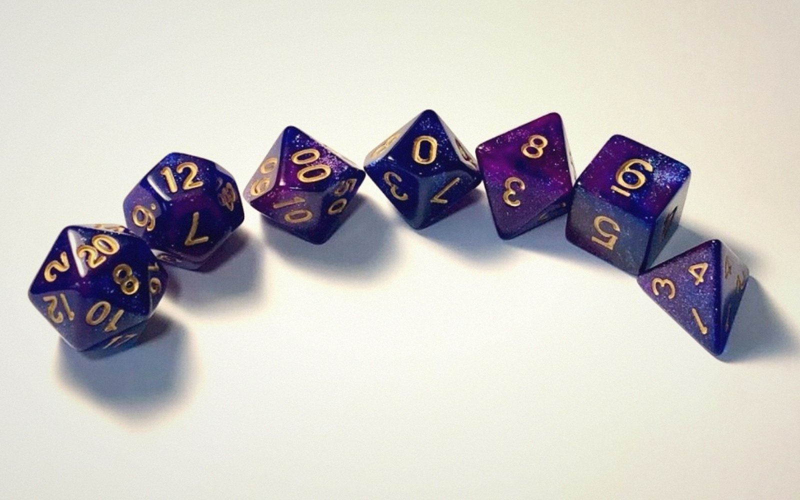 Seven polyhedral dice with gold numerals arranged in a curved line on a white surface