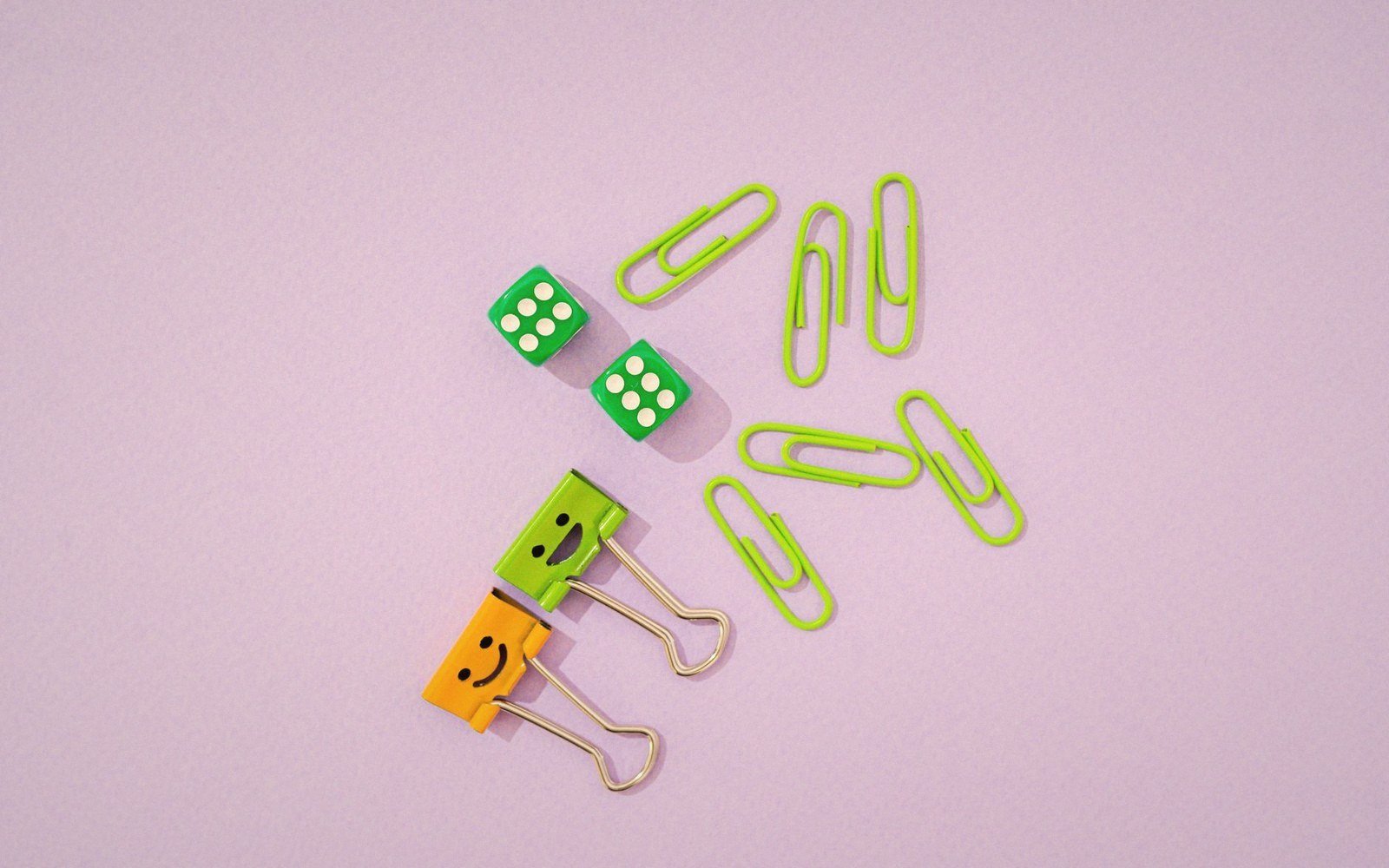 Green dice, smiley-face binder clips, and green paperclips arranged on a lilac background
