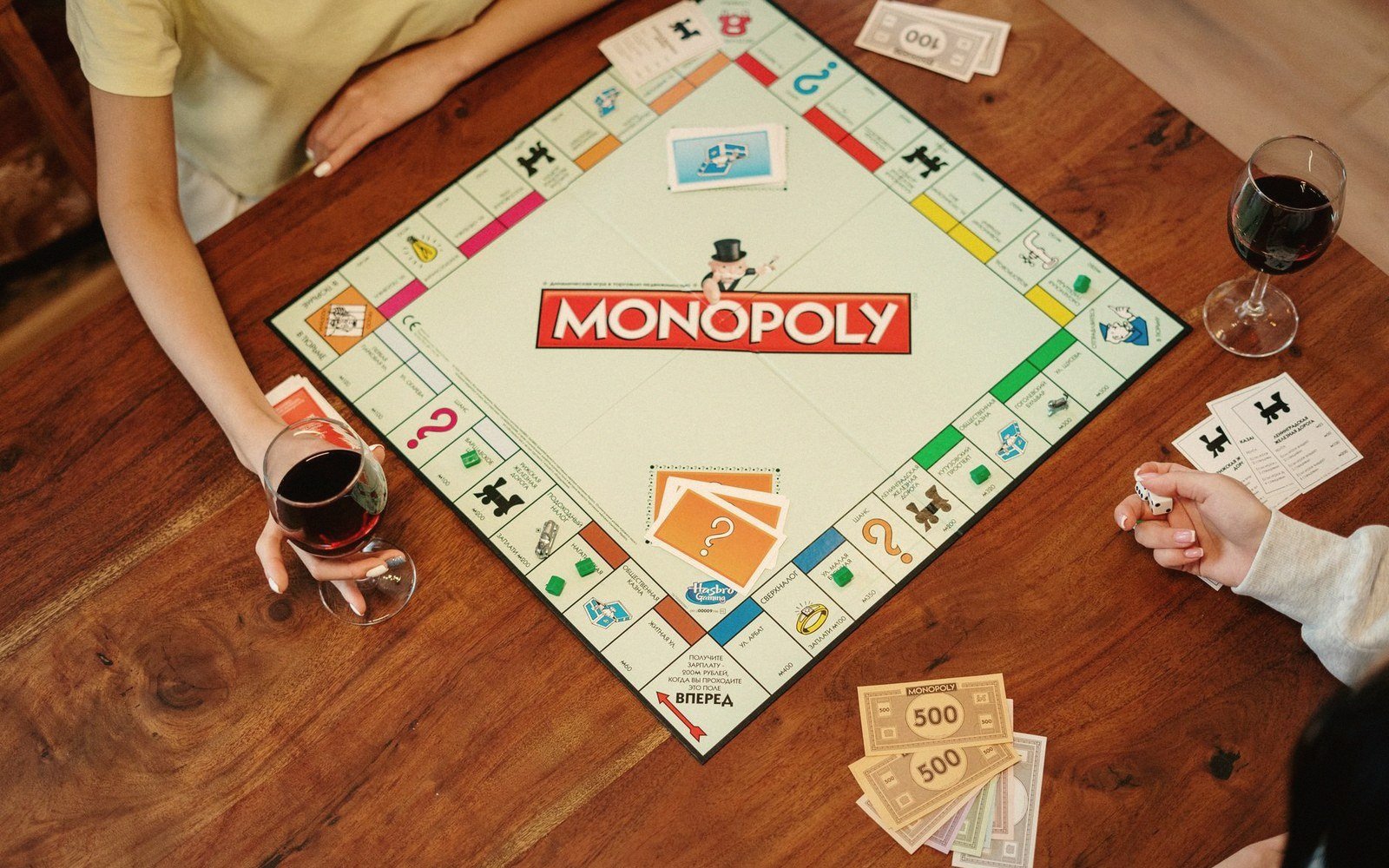 Players gather around a Monopoly board with wine glasses and cash on a wooden table