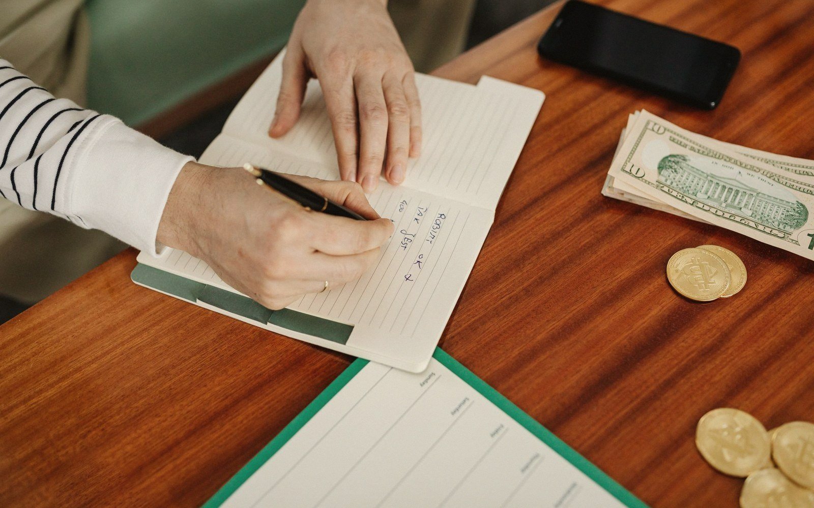 Hands writing in a notebook beside cash, gold coins and a smartphone on a desk